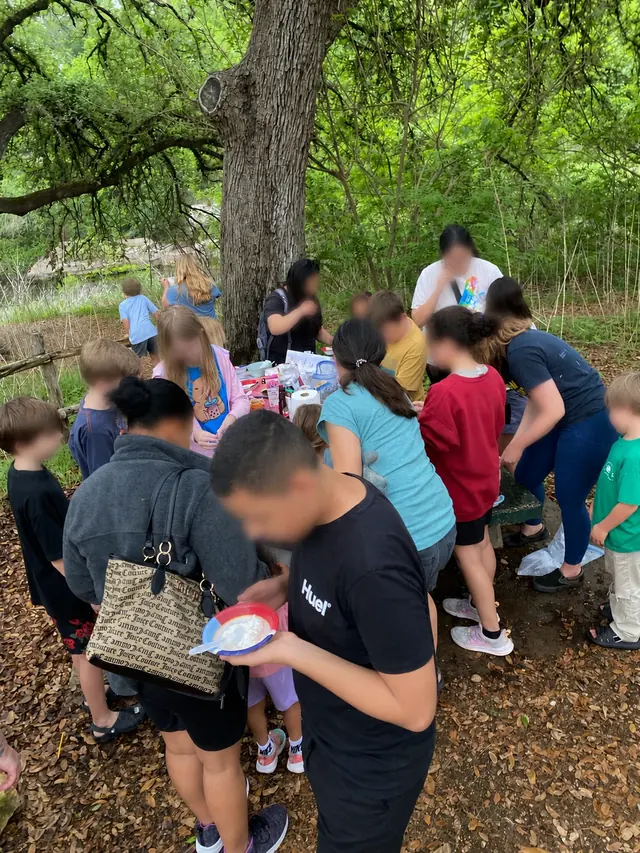 Children and adults gathered around an outdoor activity table under trees